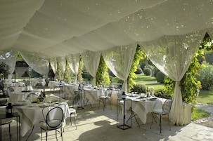 Un área de comedor al aire libre con mesas dispuestas para una comida, rodeada de telas blancas y follaje.