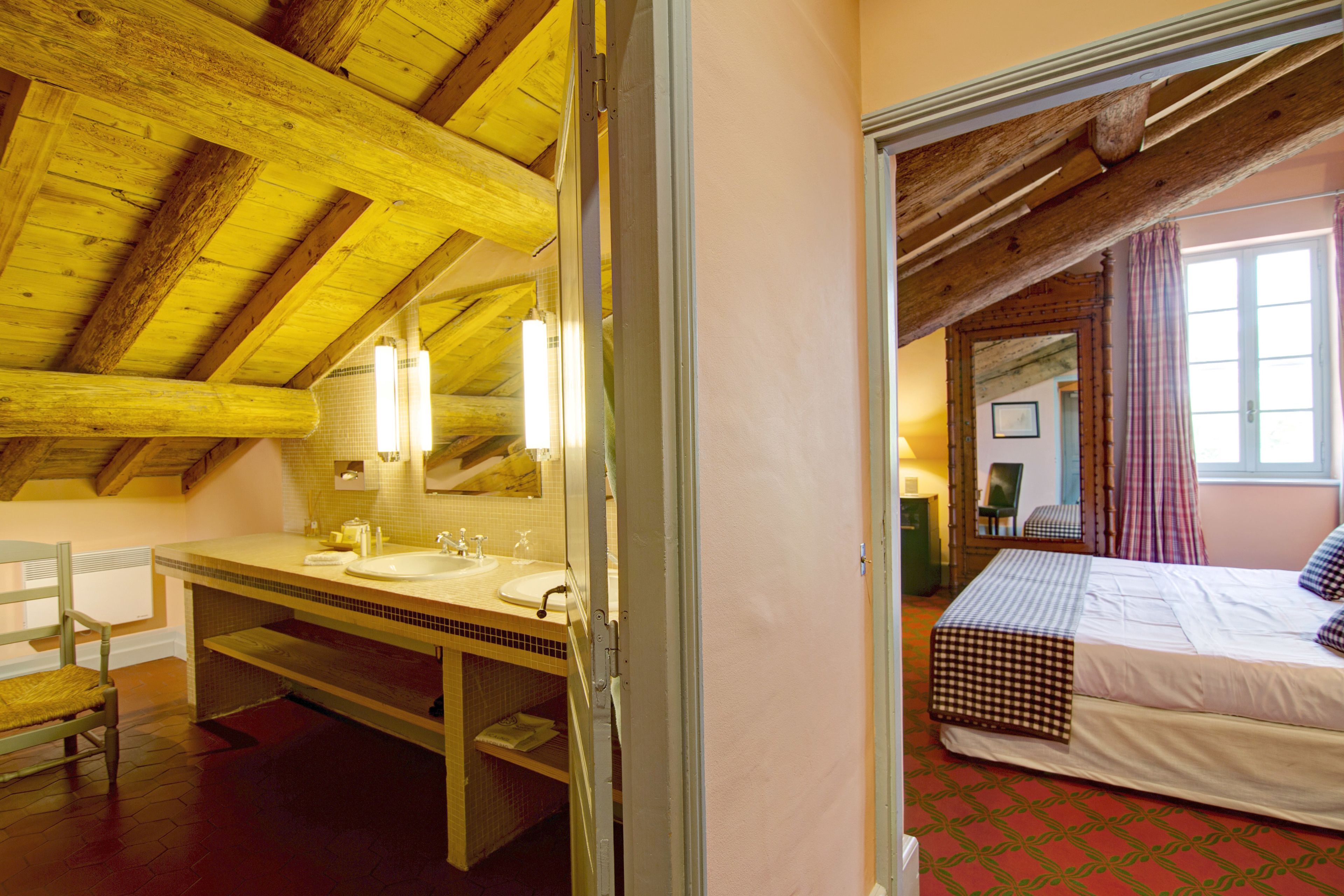 Una habitación con techo de madera, cama con colcha a cuadros, ventana grande con cortinas y baño con lavabo y espejo.