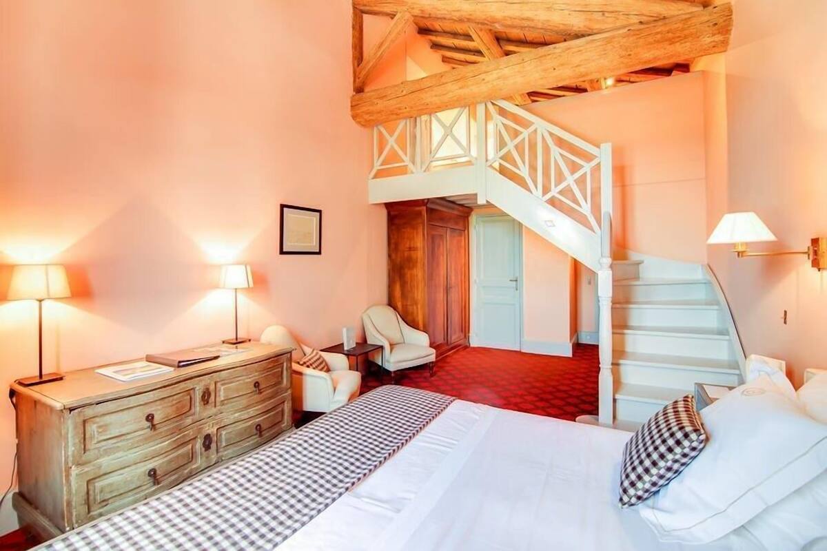 A bedroom with a bed, dresser, two chairs, a staircase, and a wooden ceiling.