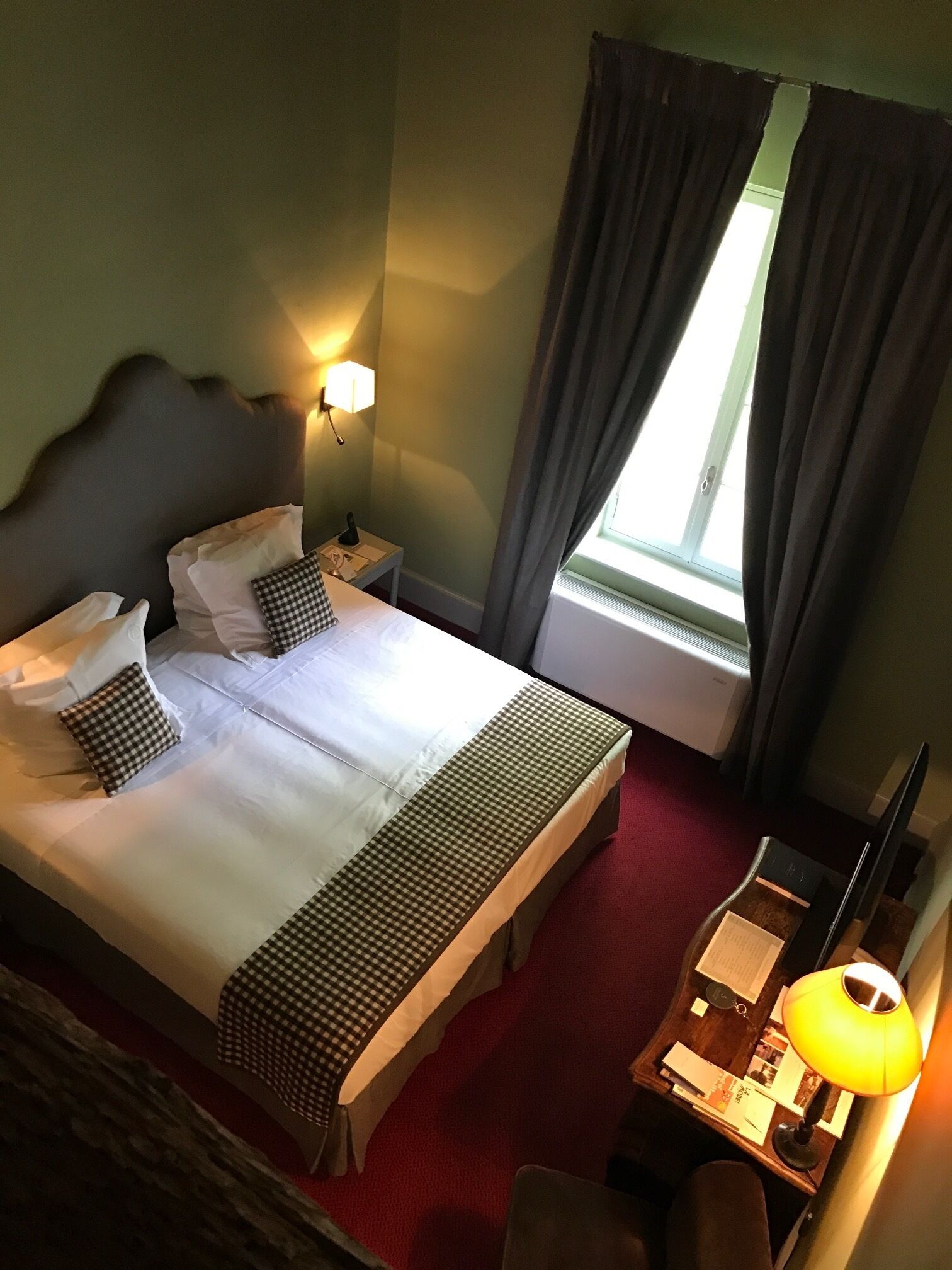 A hotel room with a large bed, a bedside table with a lamp, a desk with a chair, and a window with curtains.