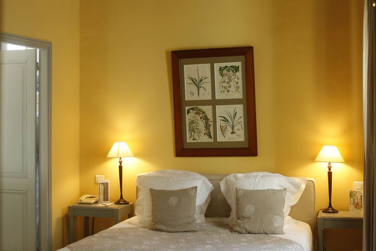 A hotel room with a bed, two bedside lamps, a ceiling fan, and a framed botanical artwork on the wall.