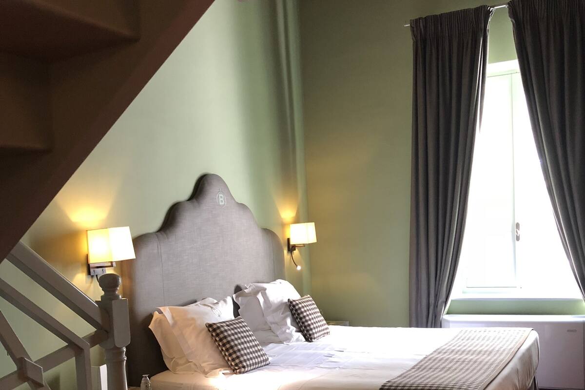 A bedroom with a bed, two bedside lamps, a window with curtains, and a staircase.