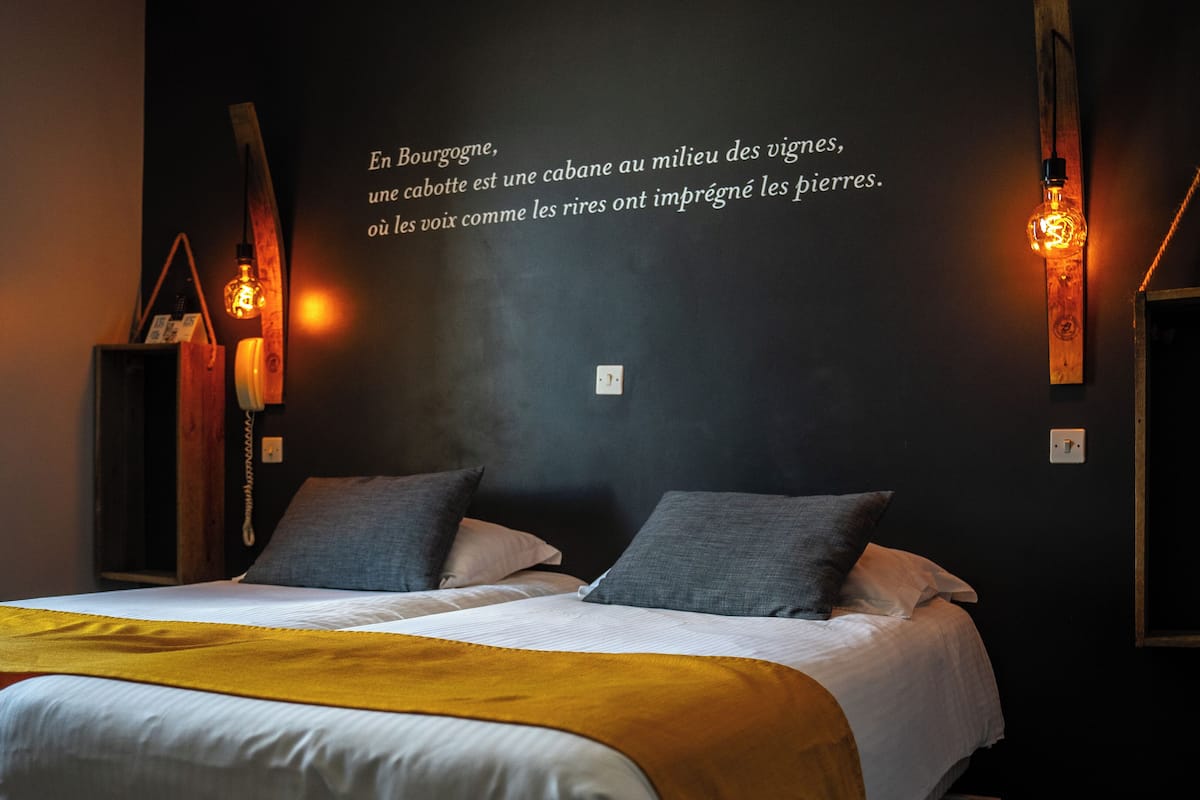 A modern hotel room with a large bed, two pillows, a yellow throw blanket, and a wall with a French quote.
