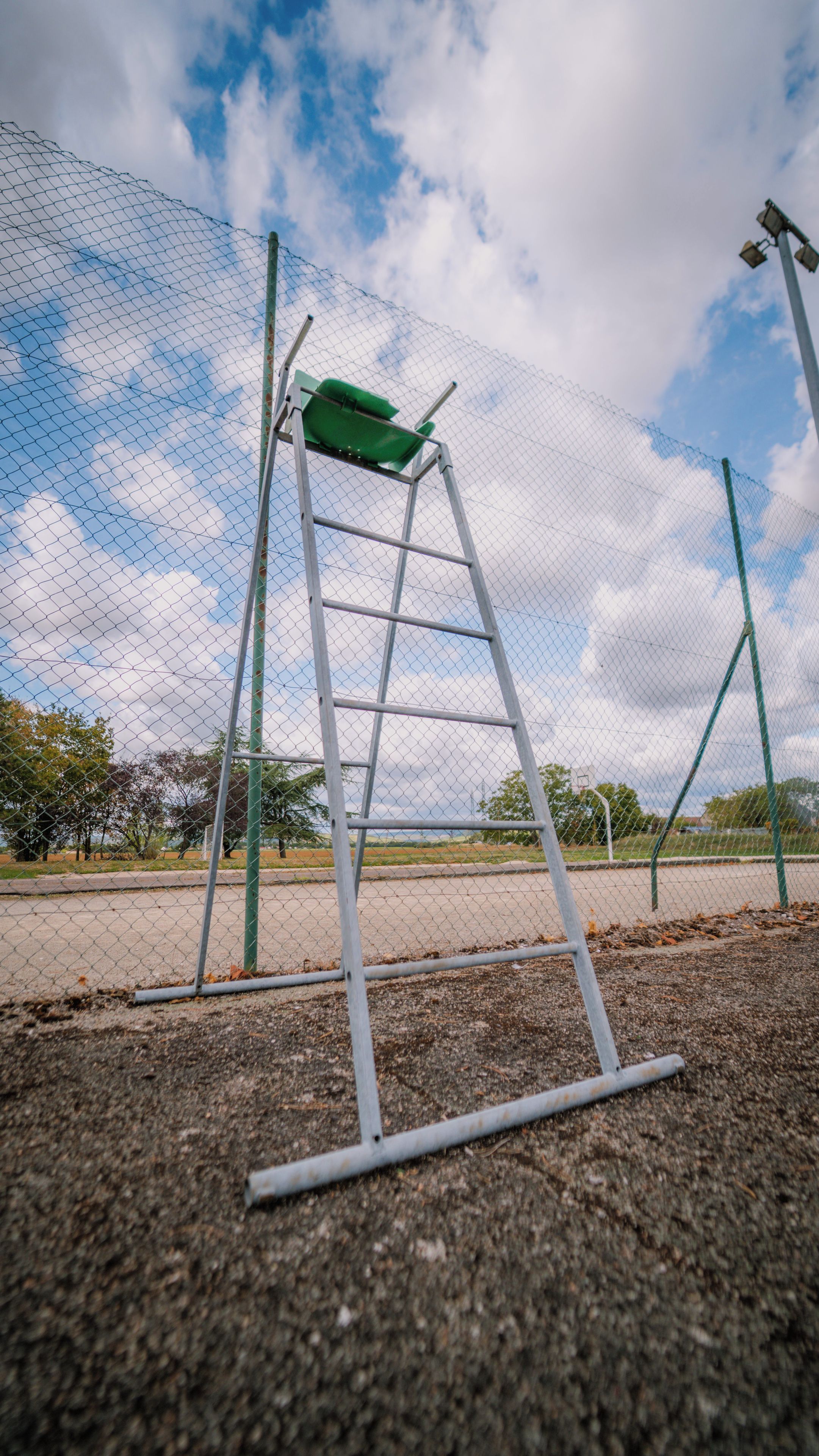 A metal ladder leaning against a chain-link fence with a green seat on top, set against a cloudy sky.