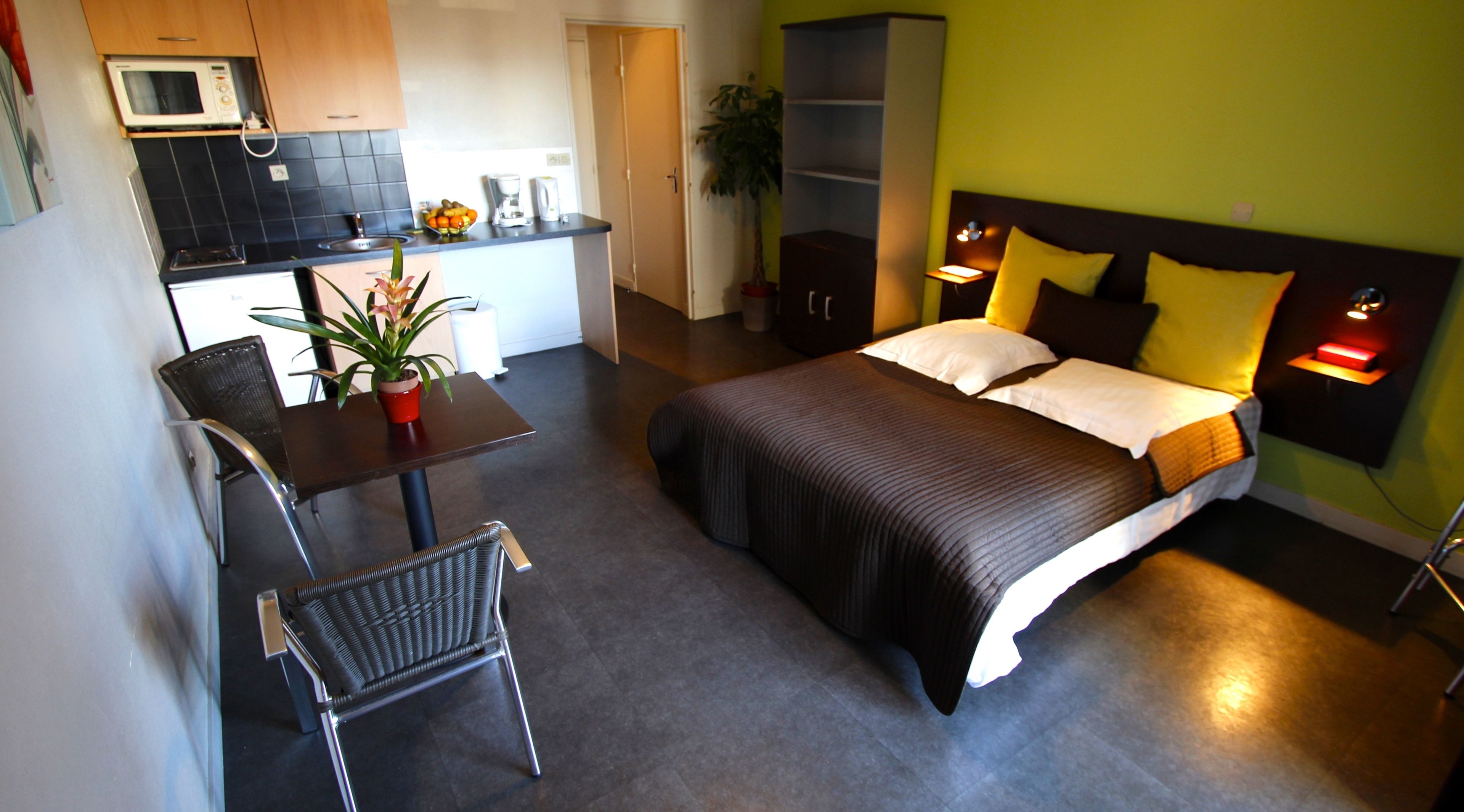A compact hotel room with a bed, bedside tables, a chair, a small kitchenette, and a potted plant.