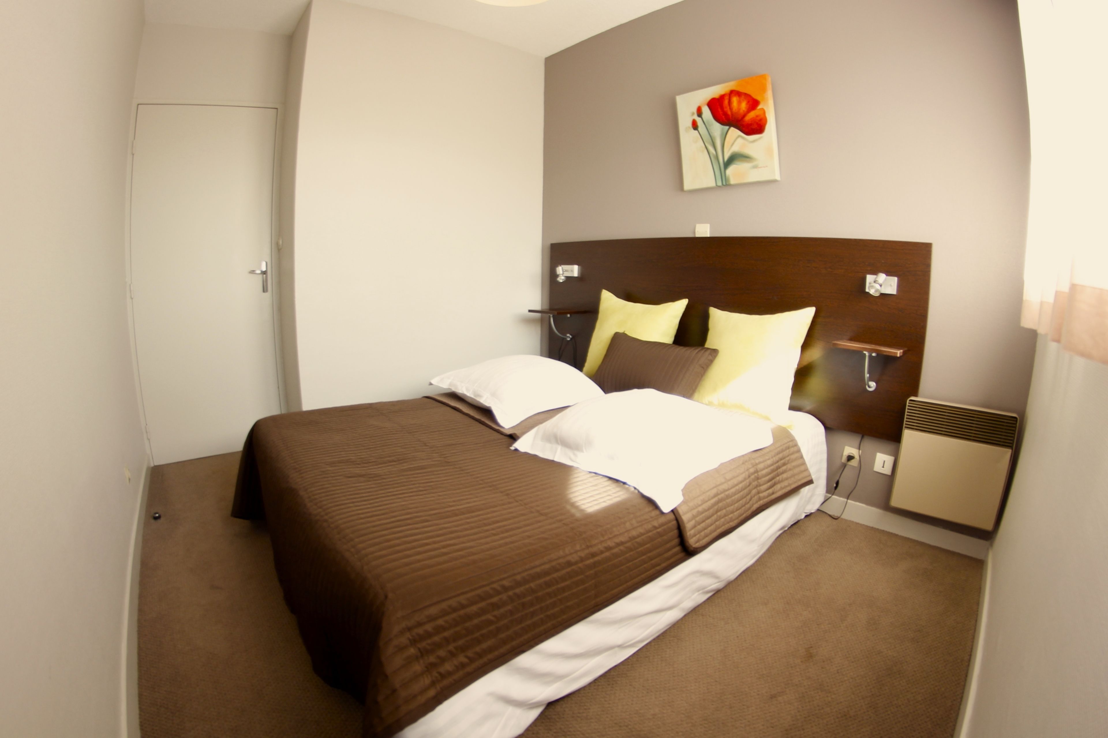 A neatly made bed with a brown and white bedspread, a wooden headboard, and a single wall-mounted nightstand with a lamp.