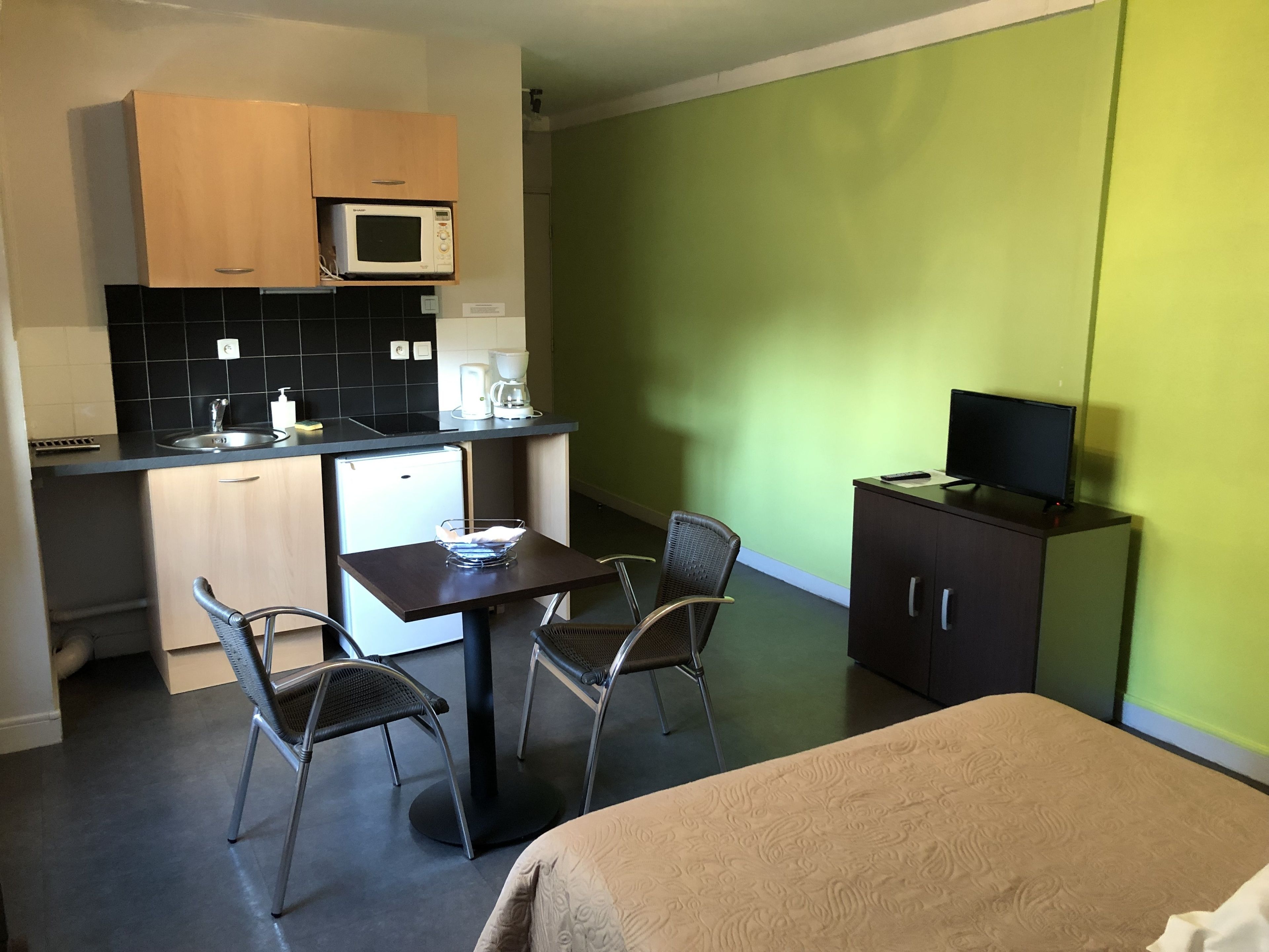 Ein kompaktes Hotelzimmer mit Küchenzeile, einem Essbereich mit zwei Stühlen, einem Bett und einem Fernseher auf einem Schrank.