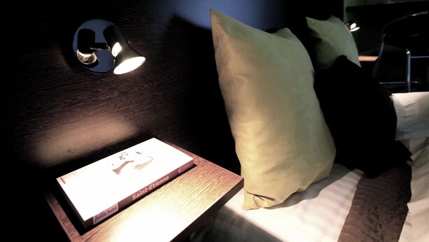 A neatly made bed with a pillow, a bedside table with a book, and a wall-mounted light fixture. - Les Palatines - Residence (Saint-Etienne)