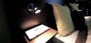 Una cama bien tendida con un almohadón, una mesita de noche con un libro, y una lámpara de pared.