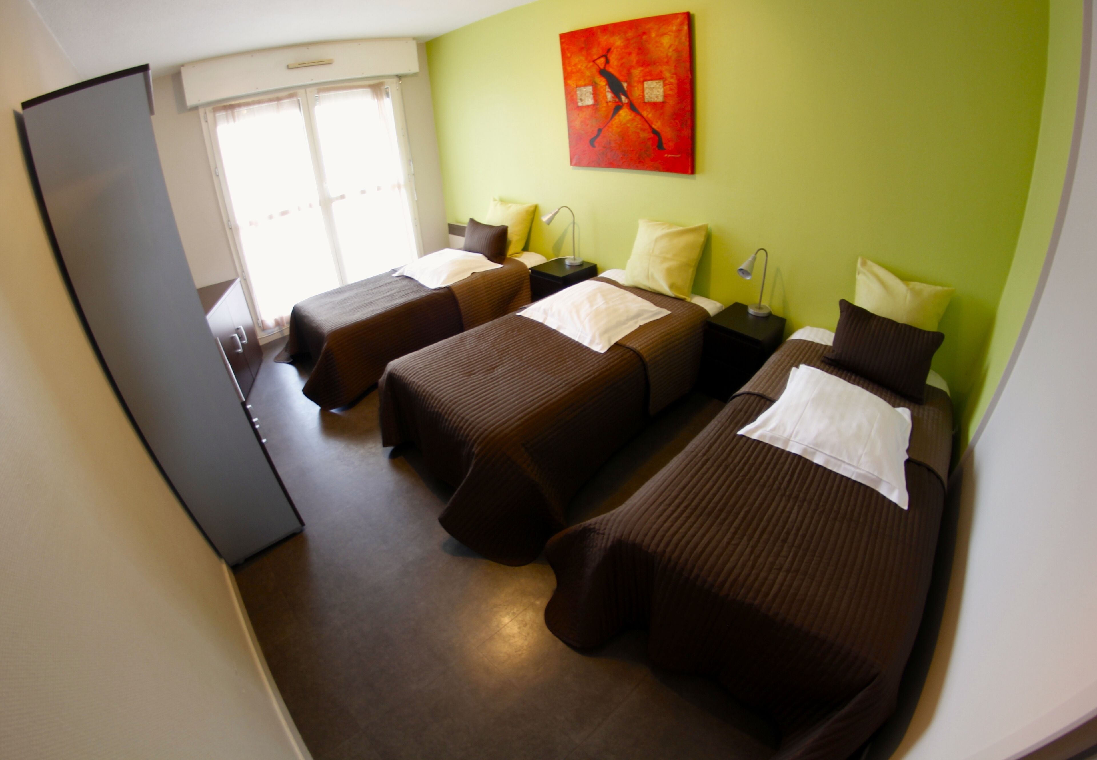 A hotel room with three single beds, a refrigerator, a window with curtains, and a painting on the wall.