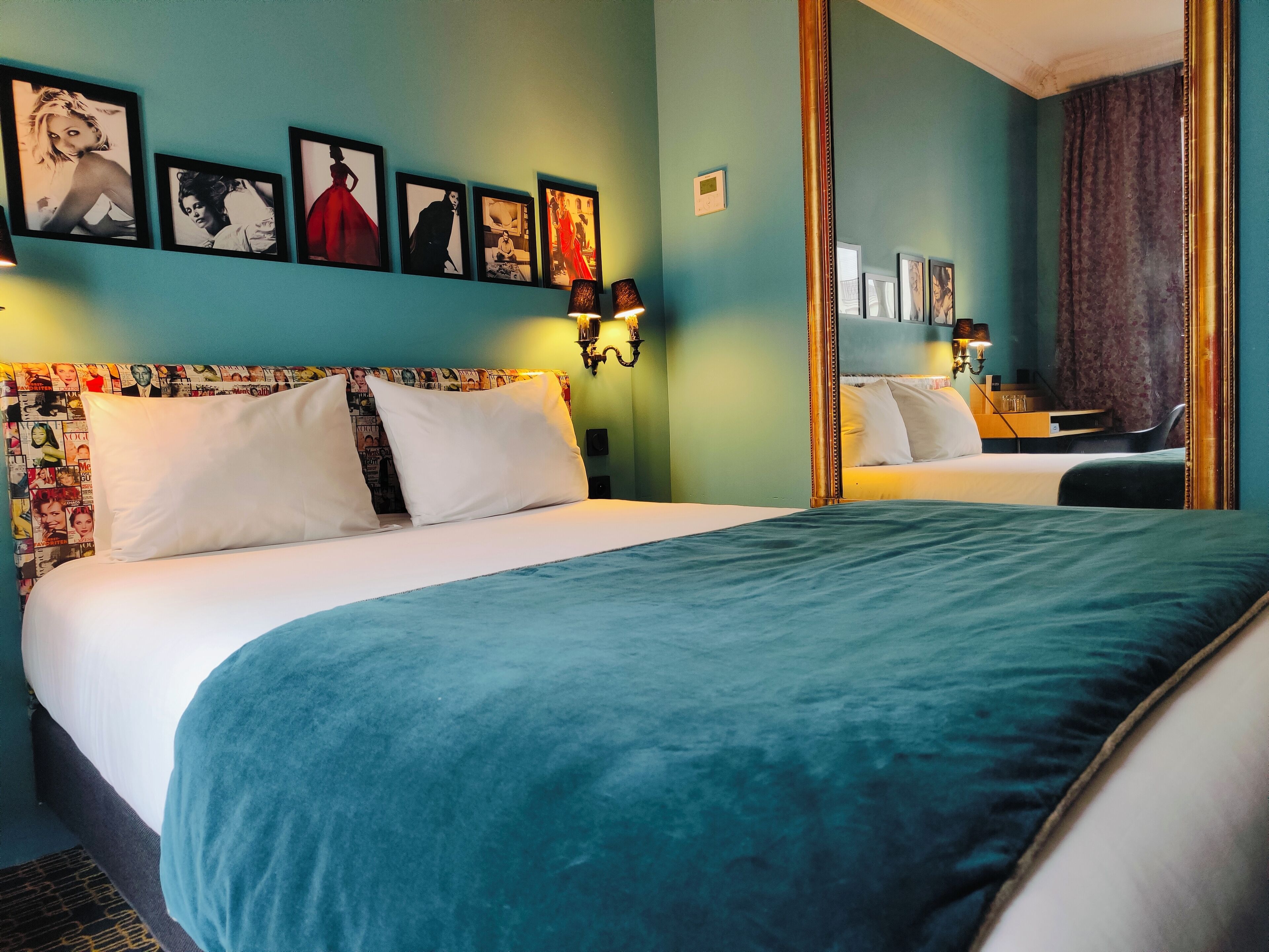 A hotel room with a large bed, a headboard with magazine covers, a mirror, and wall-mounted lights.