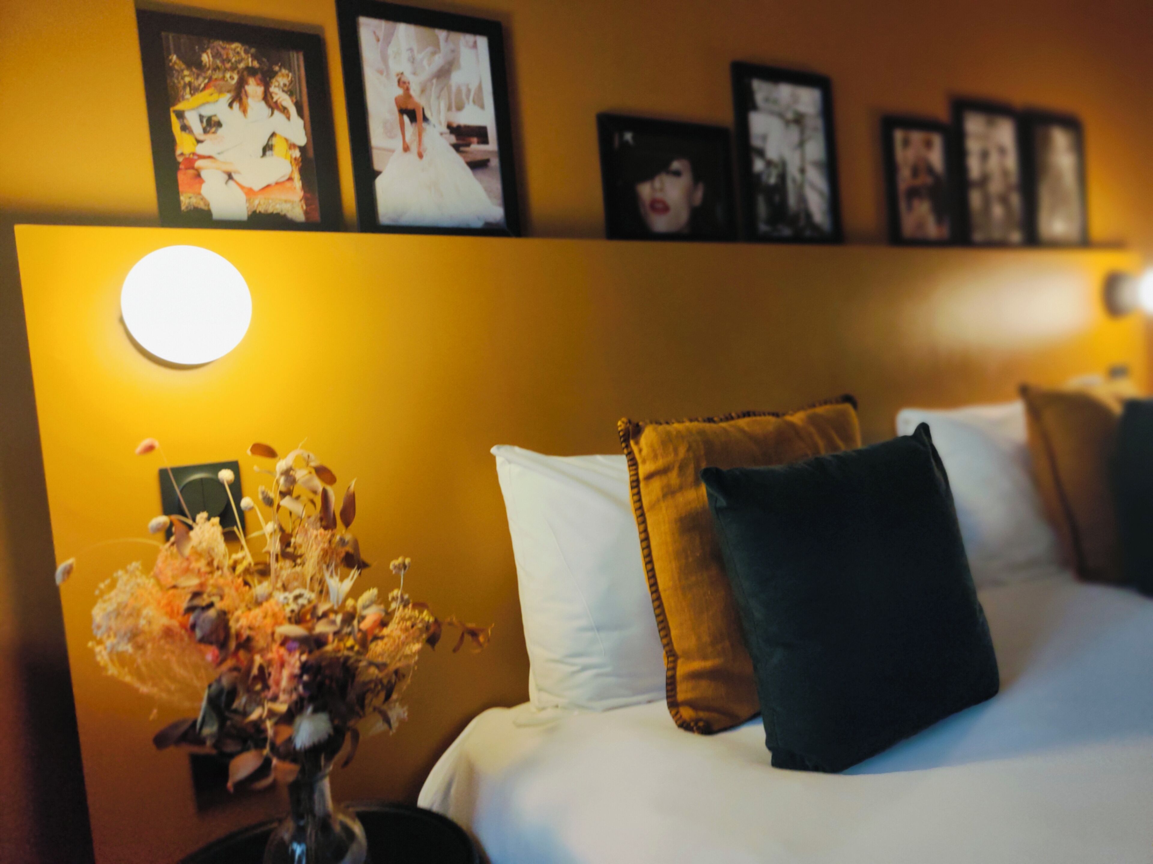 A hotel room with a bed, a nightstand with a vase of flowers, and framed pictures on the wall.