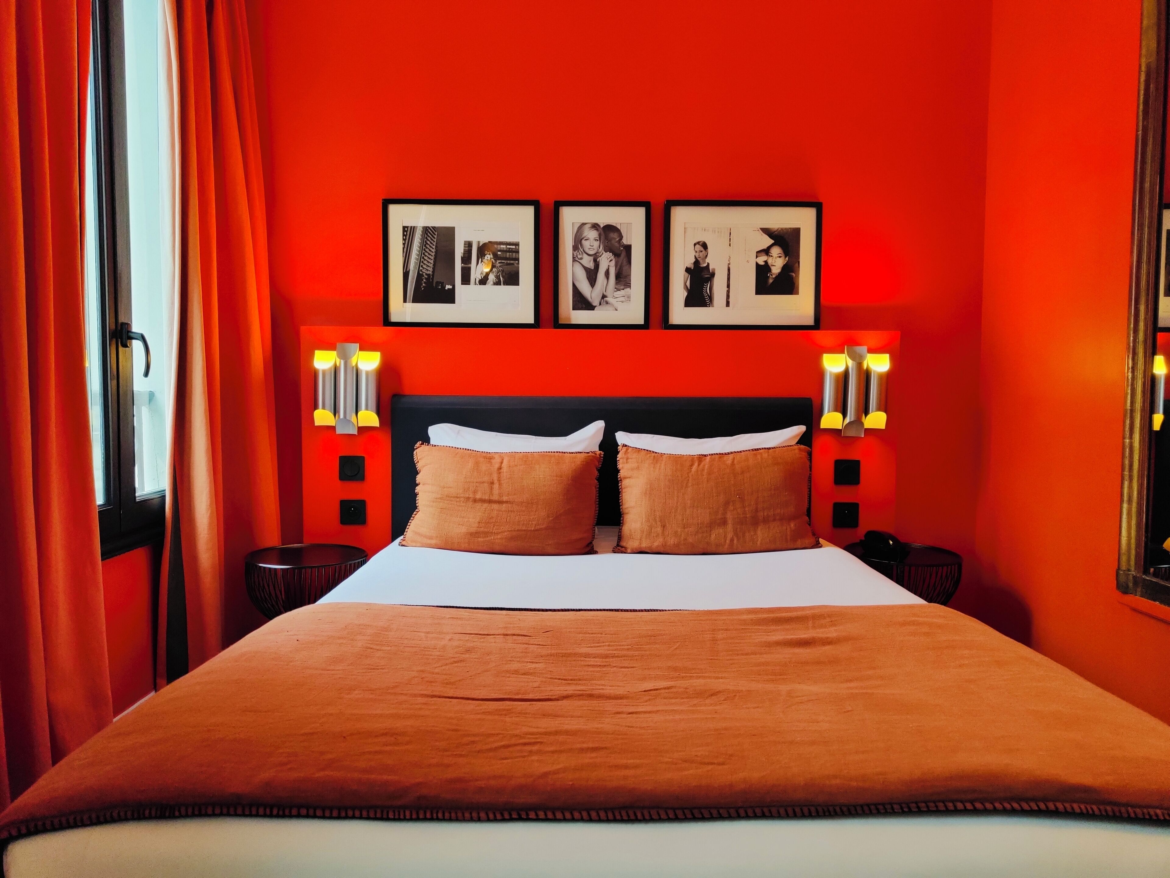 A hotel room with a red wall, a large bed with two pillows, and three framed pictures on the wall.
