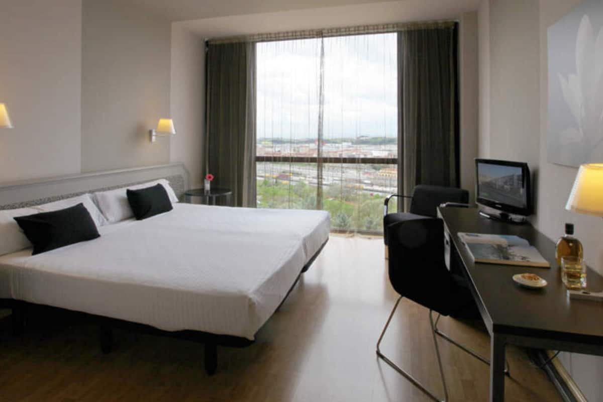 A hotel room with a large bed, a desk with a chair, a television, and a window with a view of the city.
