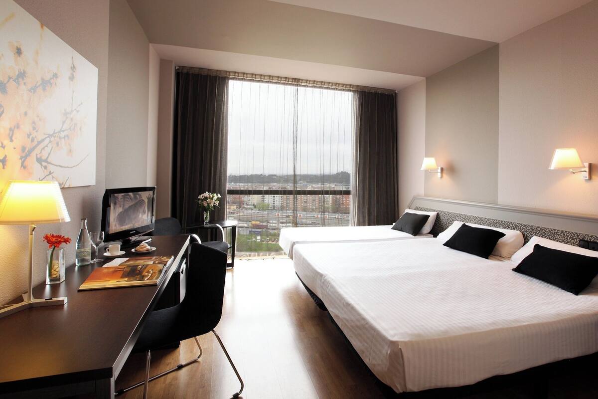 A hotel room with two beds, a desk with a computer, a chair, a lamp, and a window with a city view.