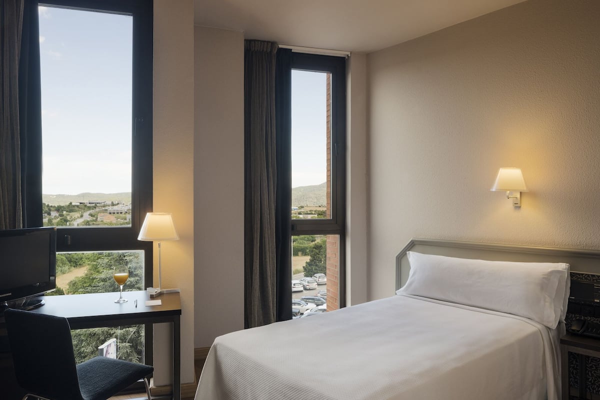 A hotel room with a bed, a desk with a lamp, and a TV. There are windows with curtains and a view of the outside.
