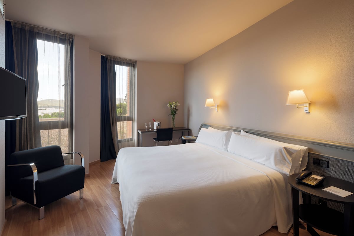 A hotel room with a large bed, a desk with a lamp, a chair, and a TV.