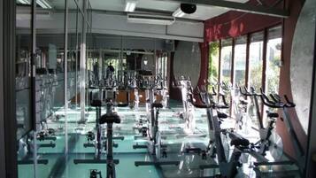 Un gimnasio con varias máquinas de ejercicio, ventanas amplias y un espejo grande.