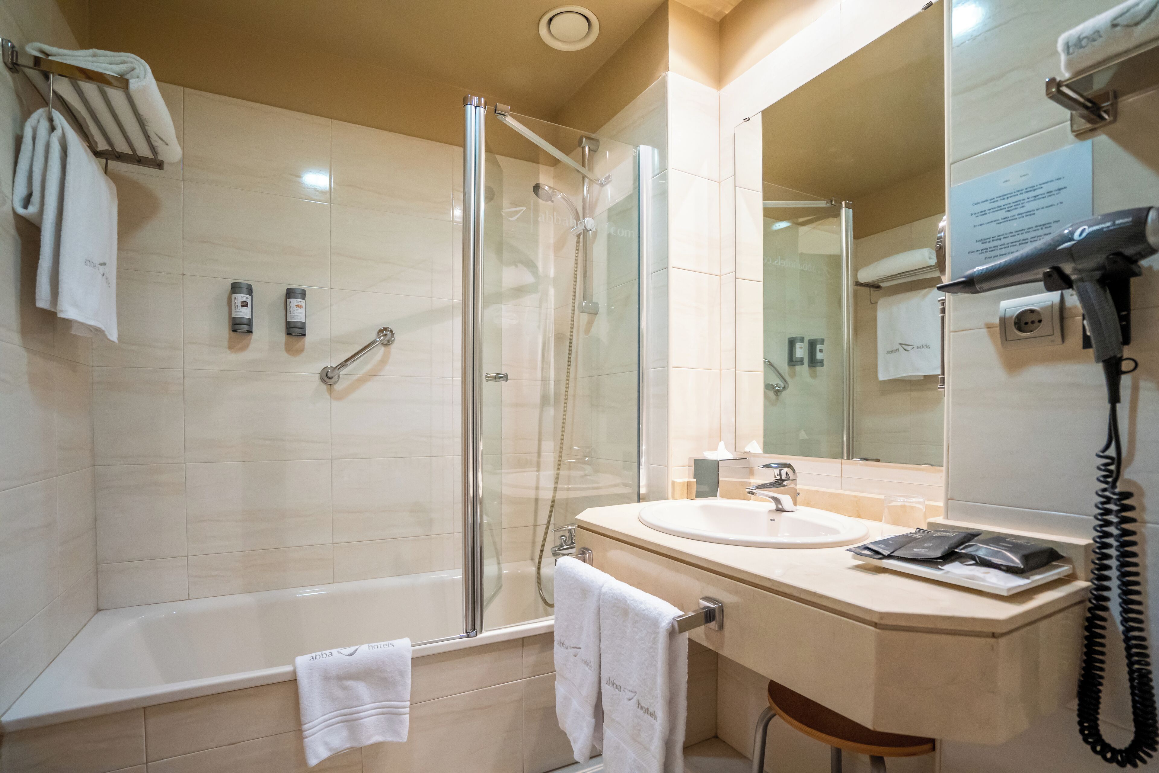 A modern bathroom with a glass-enclosed shower, a bathtub, a sink, and a hairdryer mounted on the wall.
