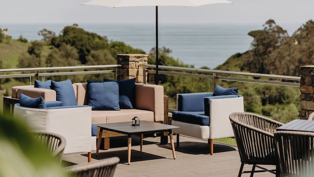 A terrace with a sofa set, a table, and chairs, overlooking a scenic view of the sea and greenery.