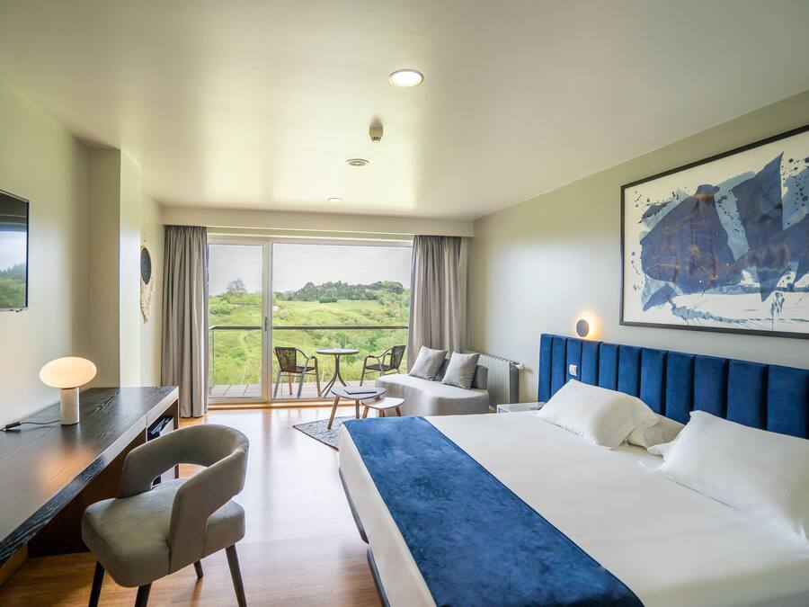 Habitación de hotel con una cama grande, un escritorio, una silla, una mesa y un balcón con vistas a zonas verdes.