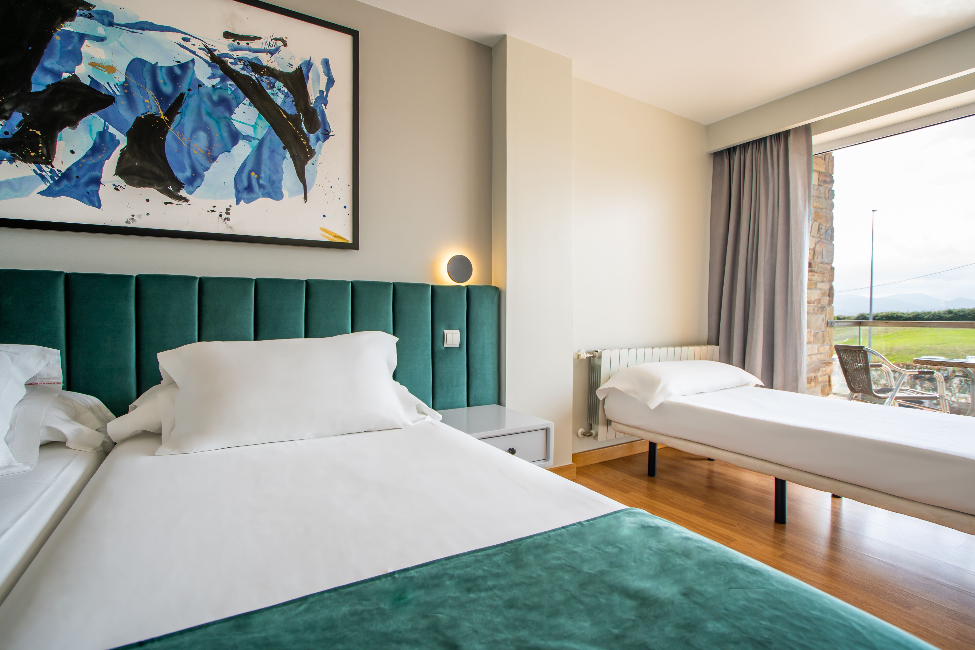 A hotel room with a large bed, a smaller bed, a painting on the wall, and a view of the outdoors through a window.
