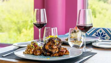 Una mesa dispuesta con platos de comida, copas de vino tinto y vistas al verdor a través de una ventana.