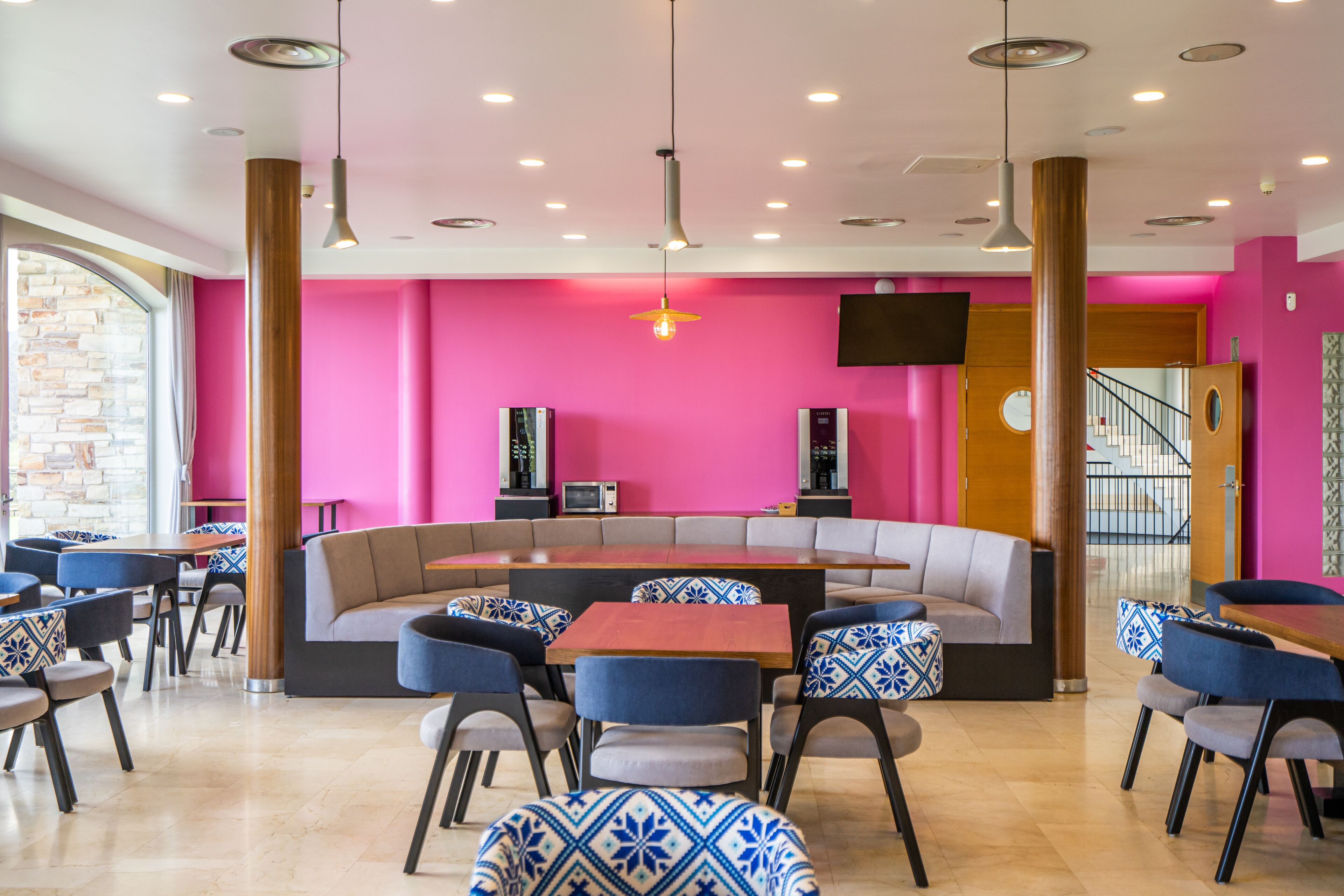 A modern cafe with a vibrant pink wall, wooden pillars, and a mix of blue and grey seating.