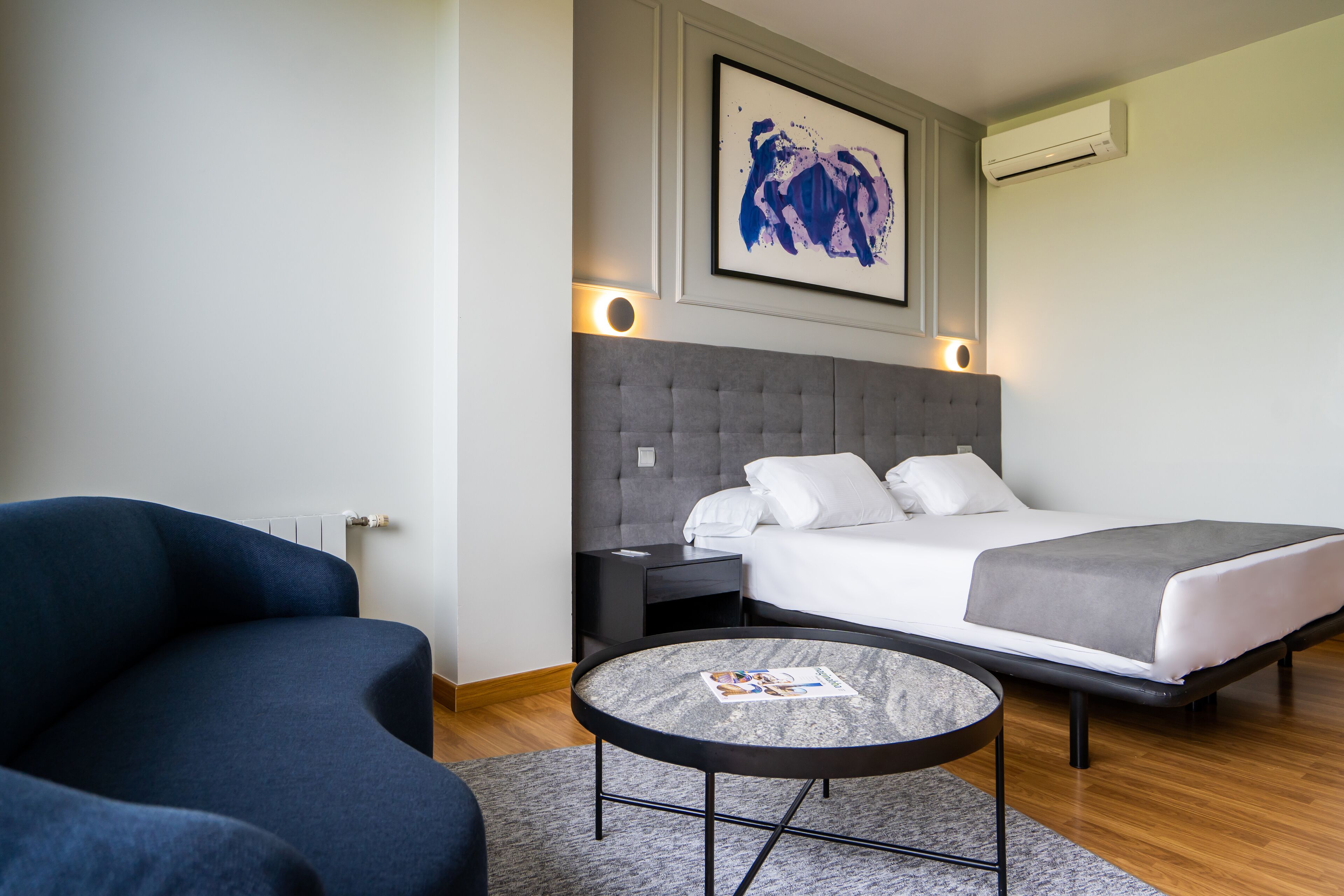 A modern hotel room with a bed, a round coffee table, a sofa, and a wall-mounted air conditioner.