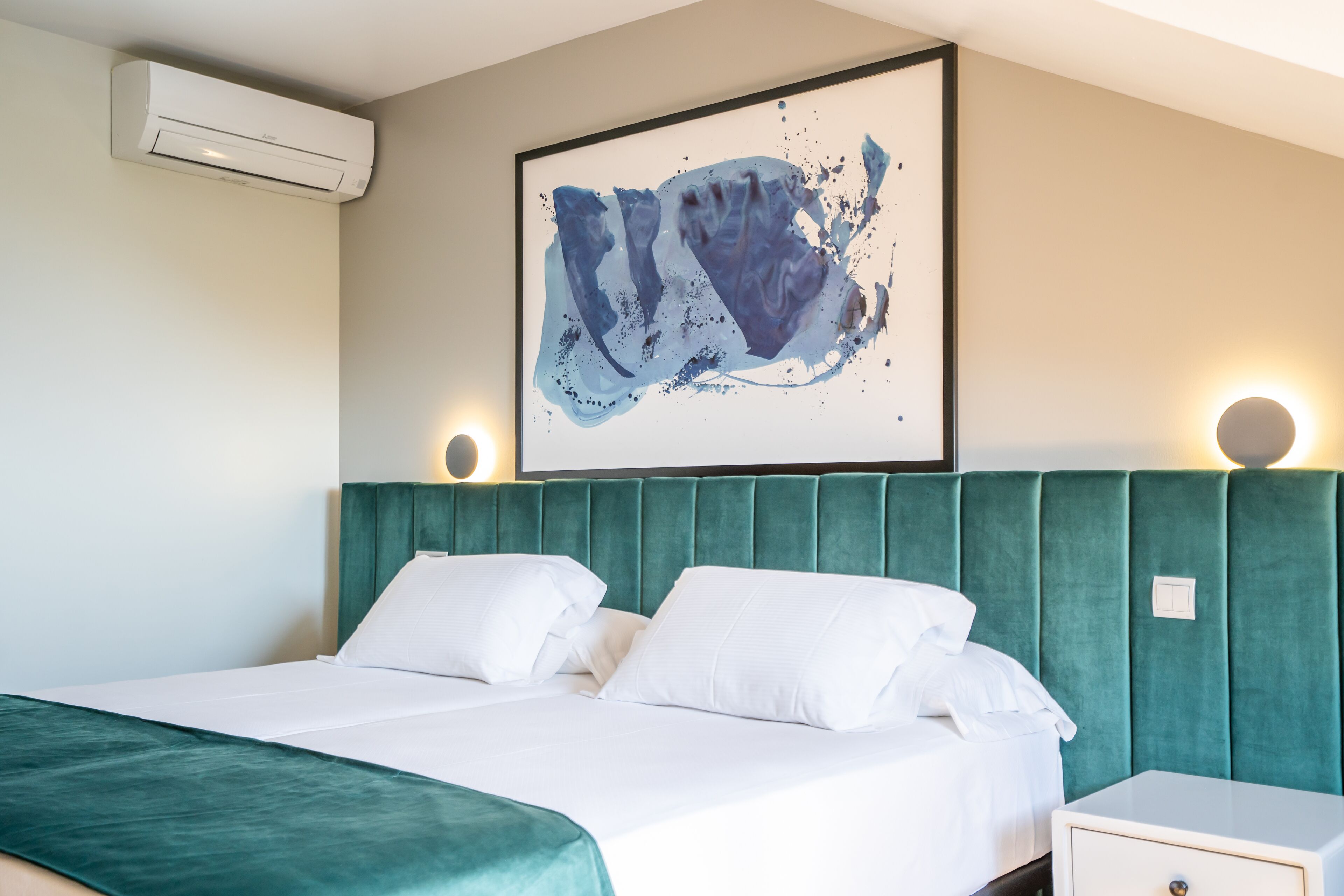 A hotel room with a bed, a green upholstered headboard, a framed abstract painting, and an air conditioning unit.