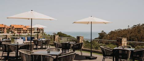 An outdoor patio with wicker chairs and tables, white umbrellas, and a view of the ocean and buildings in the background.