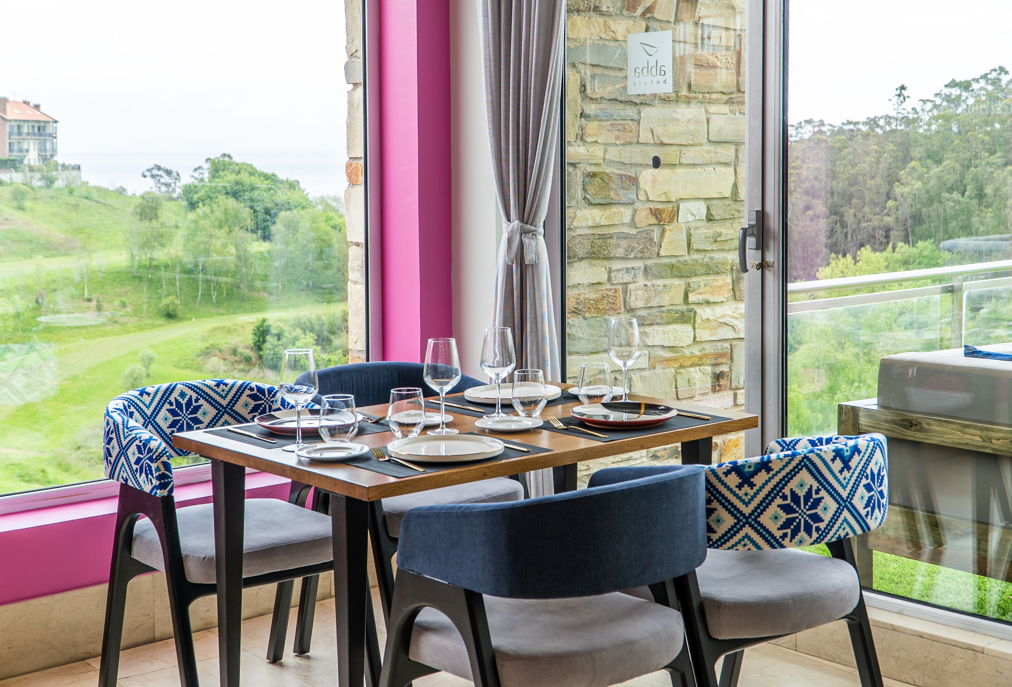 Un comedor con una mesa dispuesta para cuatro, sillas con cojines estampados, una pared de piedra y vistas a un paisaje verde.