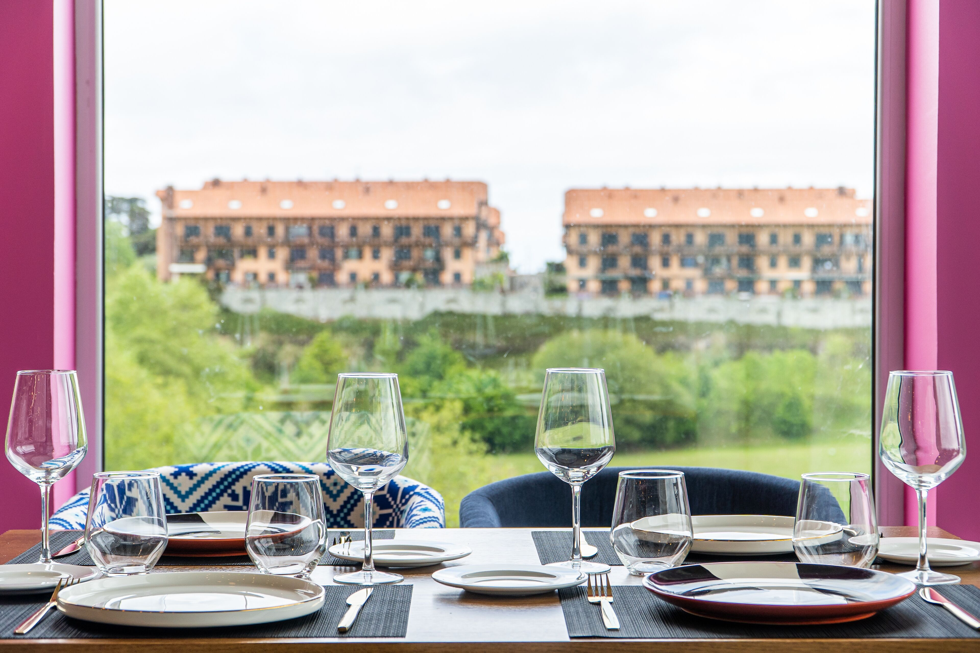 Une table dressée pour le dîner, avec vue sur les bâtiments et la verdure, par une fenêtre.