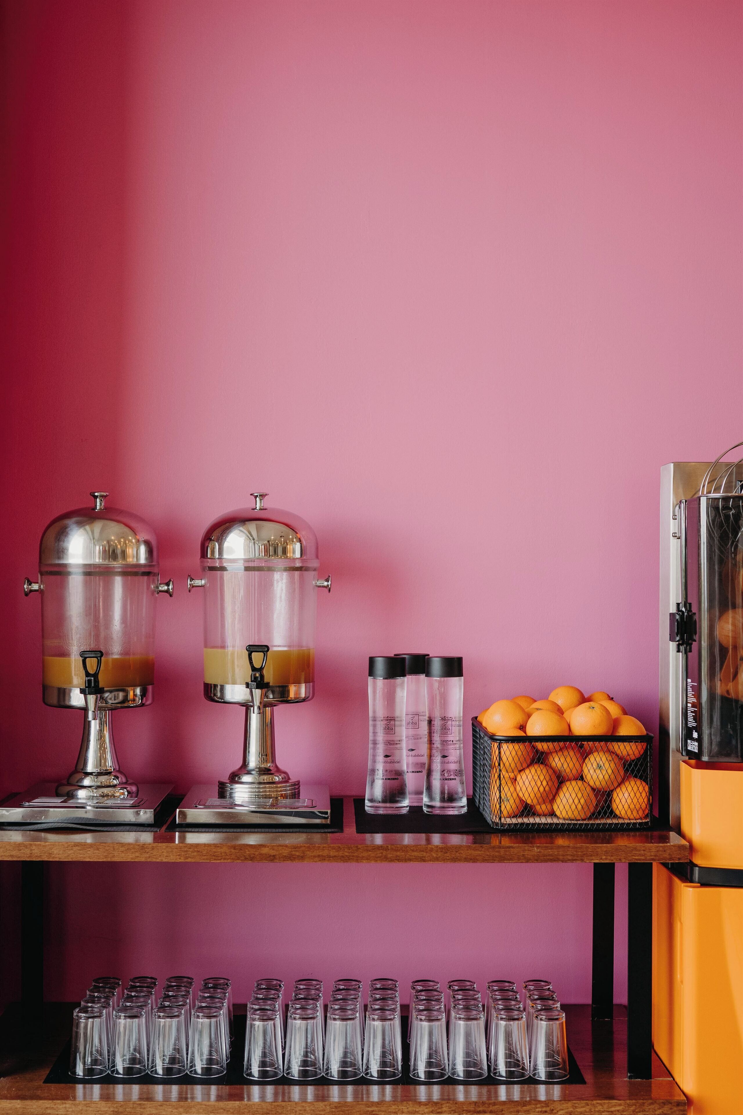 A beverage station with two dispensers, a basket of oranges, and a row of glasses on a wooden table against a pink wall.