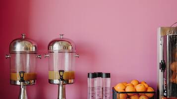 A beverage station with two dispensers, a basket of oranges, and a row of glasses on a wooden table against a pink wall.
