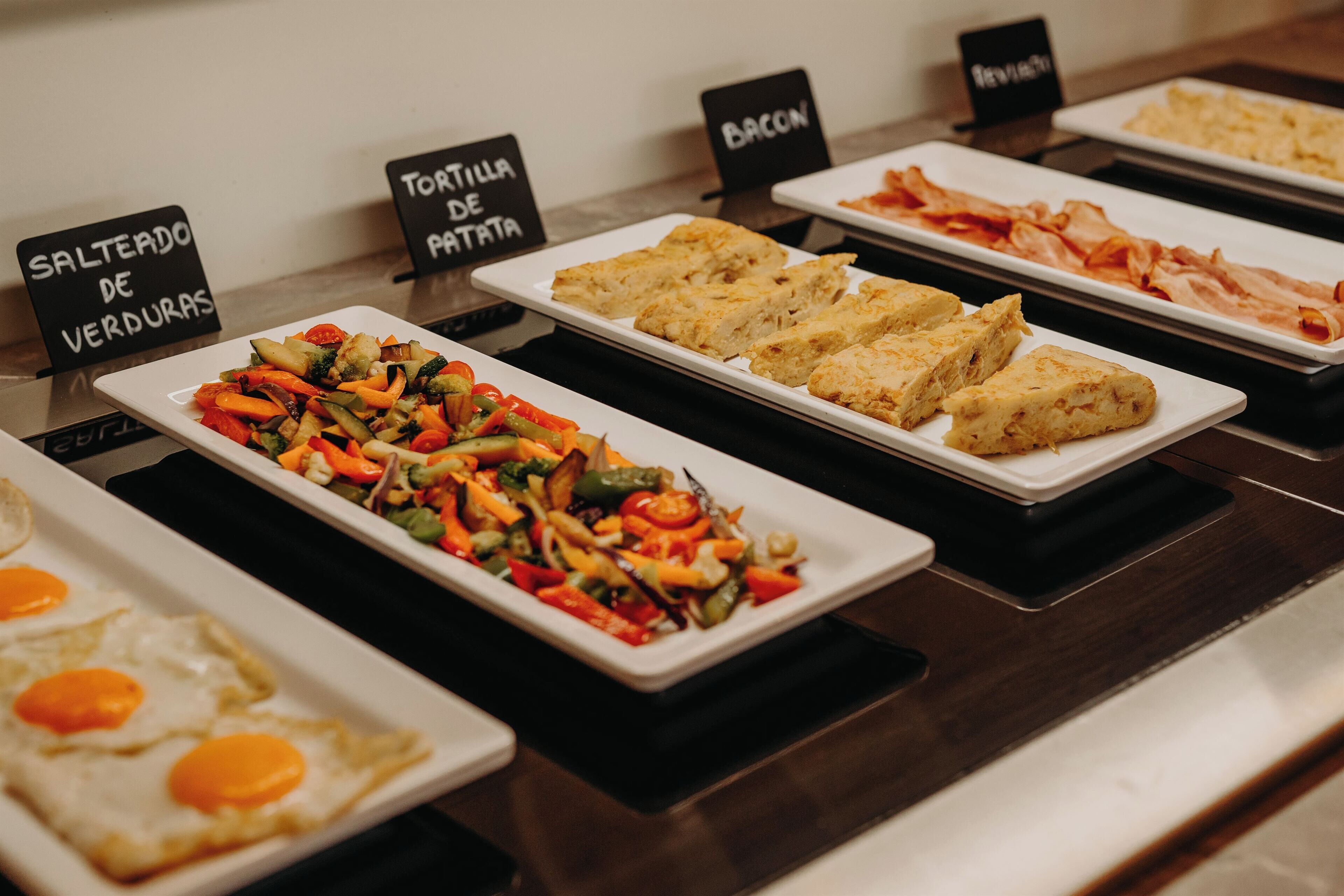 A buffet with labeled dishes including 'Salteado de Verduras', 'Tortilla de Patatas', and 'Bacon'.