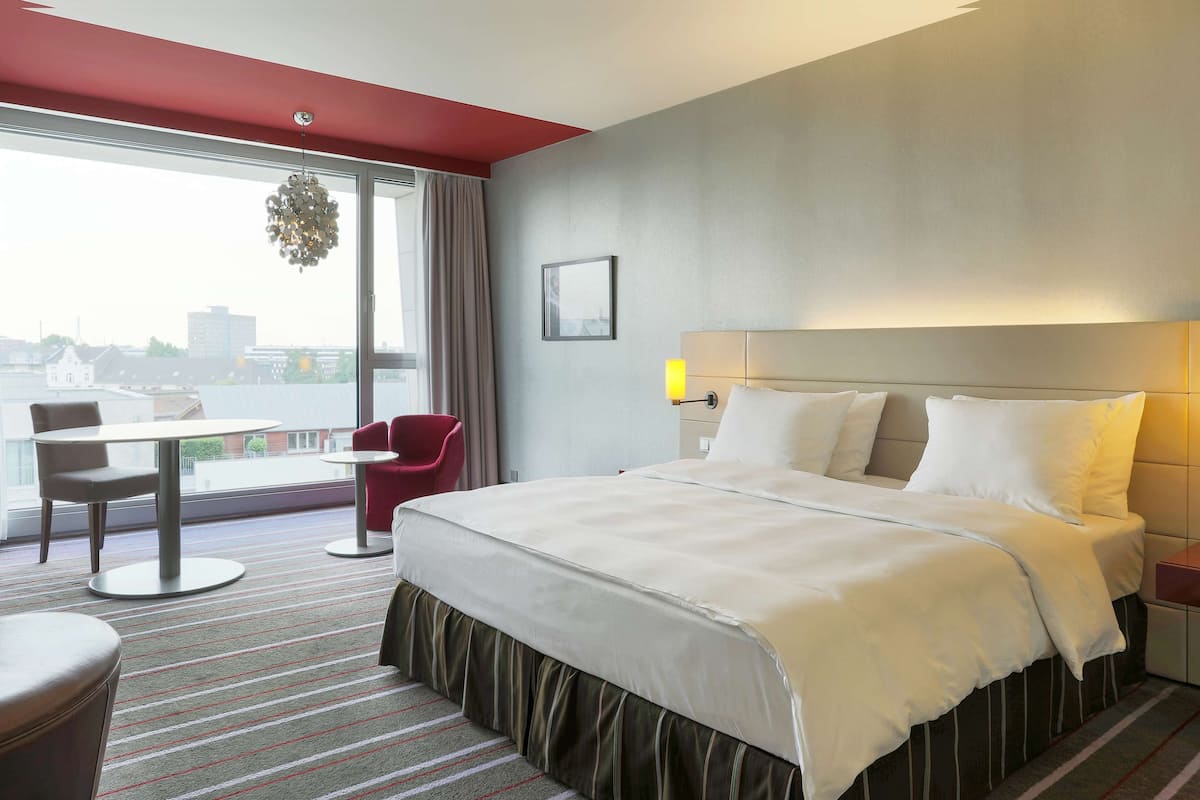 A hotel room with a bed, a desk, a chair, a window with a view, and a red accent wall.