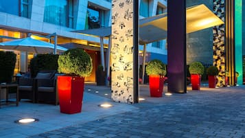 Un espacio al aire libre con mobiliario, plantas en macetas y fachada de edificio moderna.