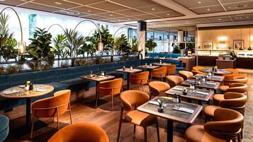 Un restaurante moderno con diferentes opciones de asientos, incluyendo mesas y sofás, variedad de plantas y vistas al exterior.