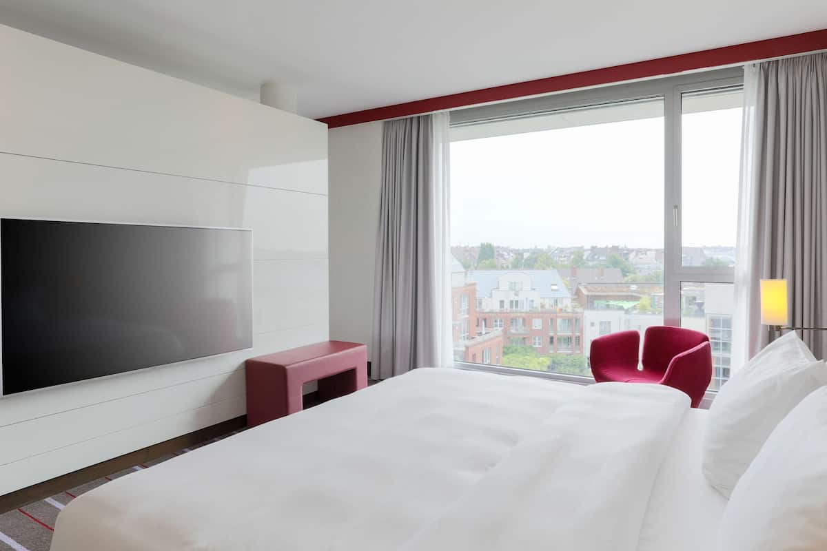 A hotel room with a large bed, a flat-screen TV, a red chair, and a view of buildings through the window.