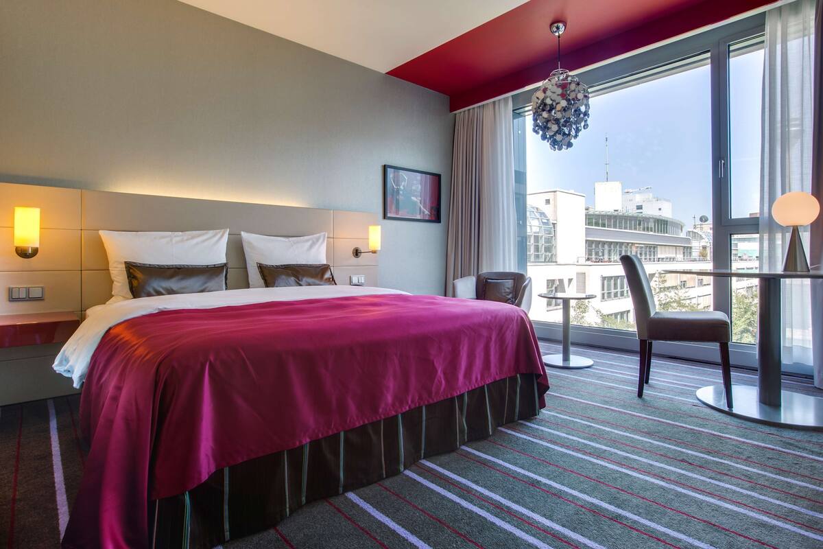 A hotel room with a large bed, a chair, a table, and a city view.