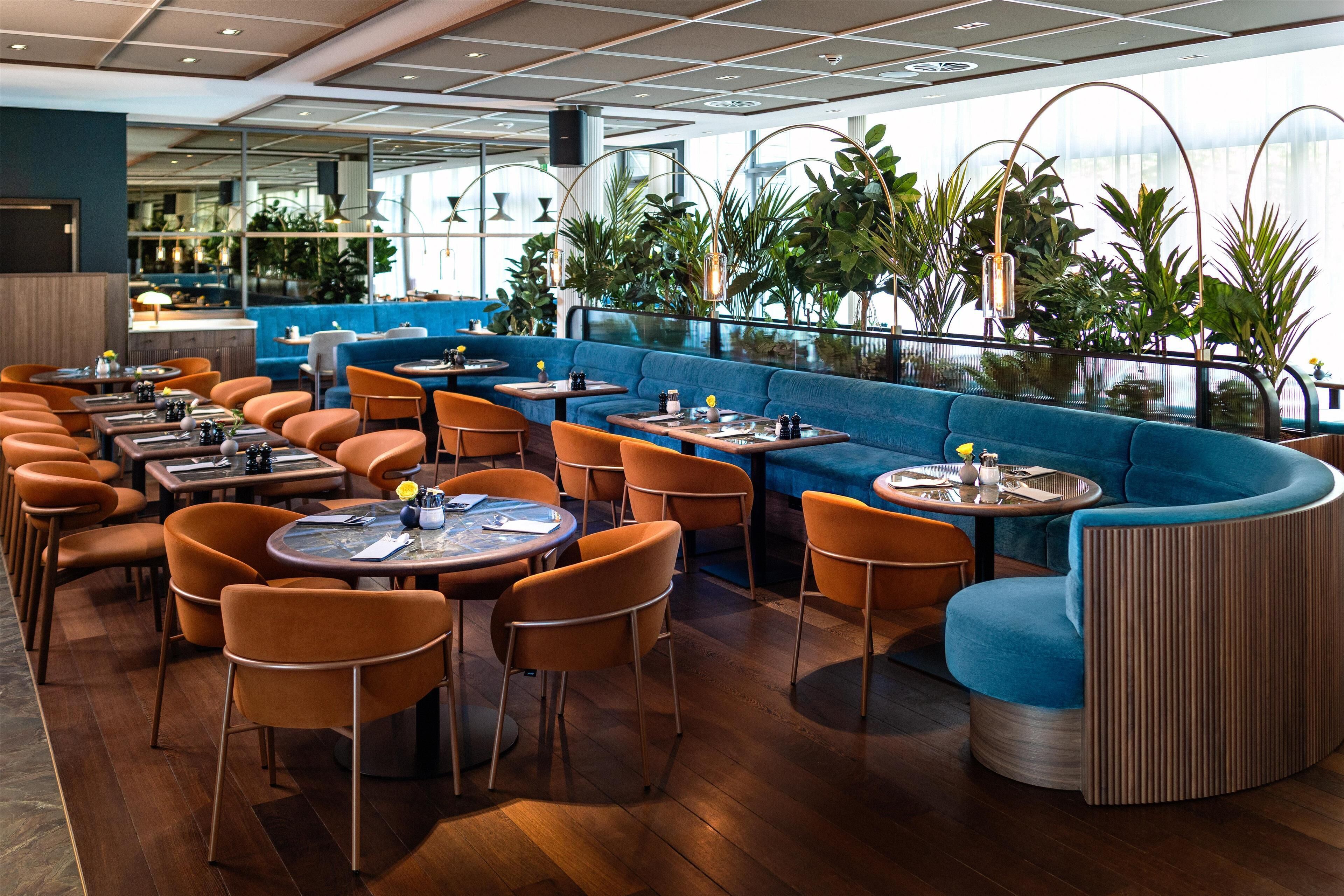 Un restaurante moderno con mesas redondas y sillas color naranja, una zona de estar azul y ventanas amplias con plantas.
