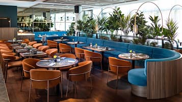 Un restaurante moderno con mesas redondas y sillas color naranja, una zona de estar azul y ventanas amplias con plantas.