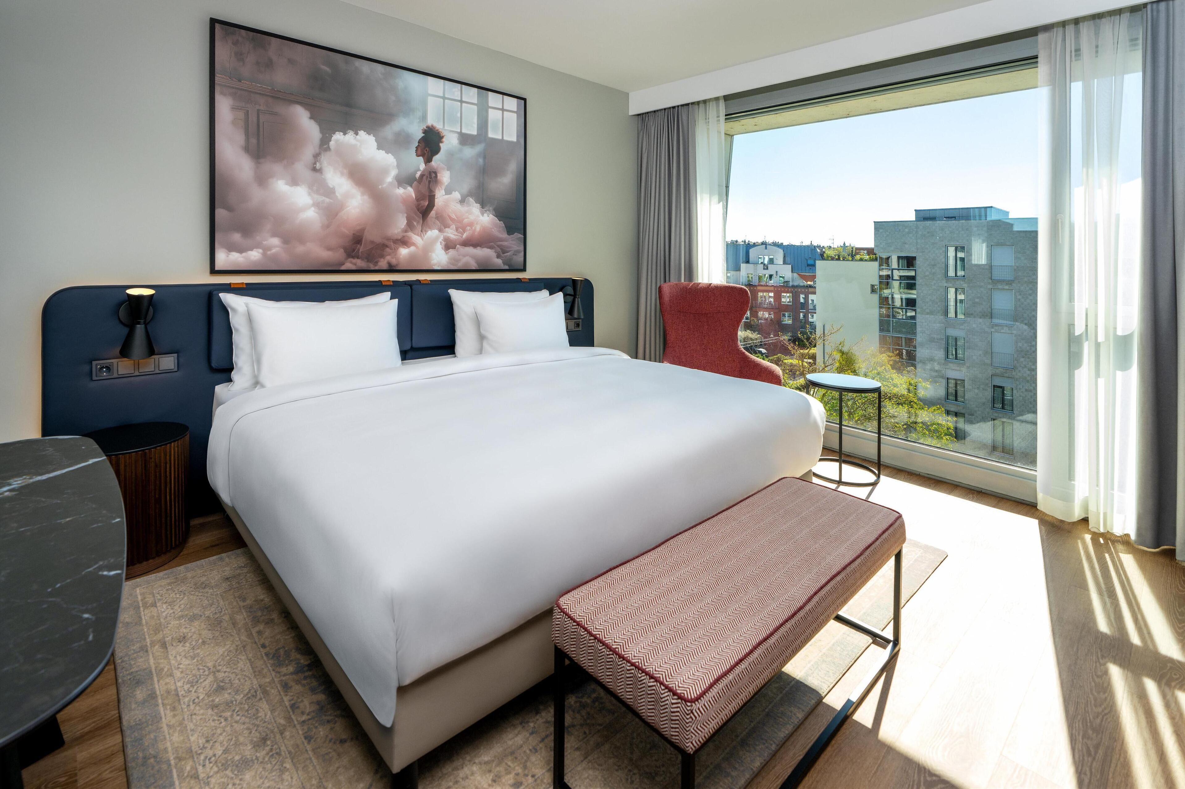 Et moderne hotelværelse med en stor seng, et sengebord, en stol og udsigt over byen gennem et stort vindue.