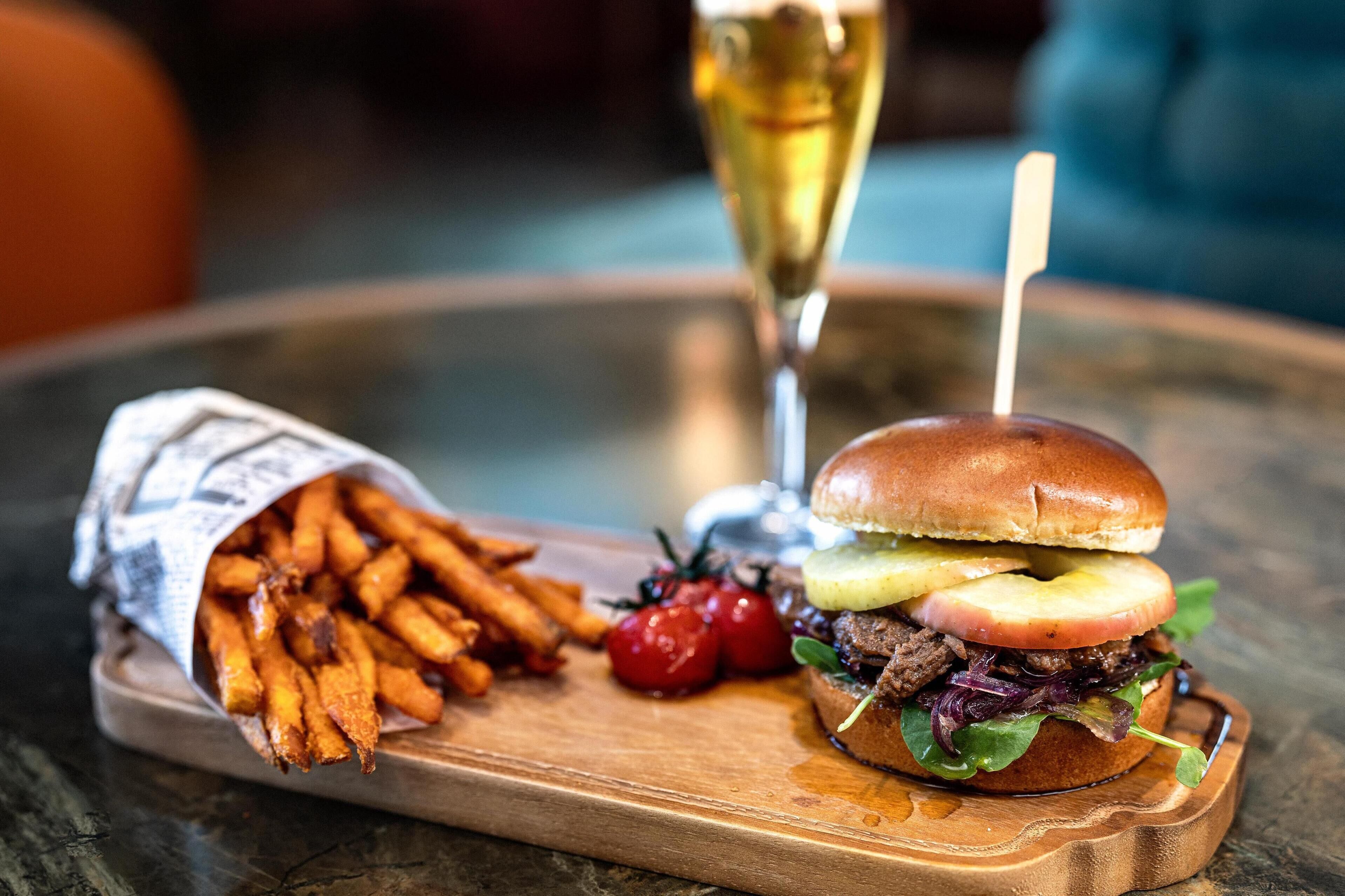 A meal consisting of a burger with lettuce, tomato, and a bun, accompanied by sweet potato fries and a glass of white wine on a wooden board.