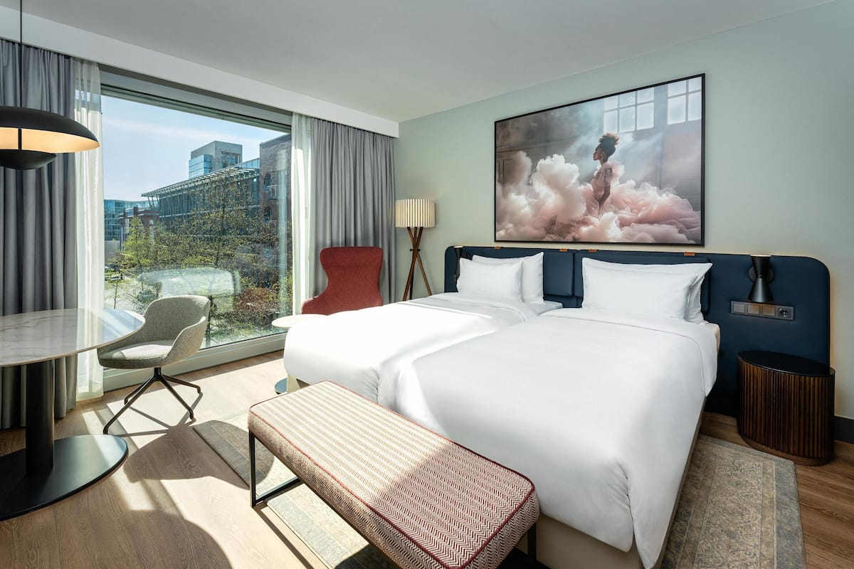 A hotel room with a large bed, a desk, a chair, a bench, a lamp, and a painting.