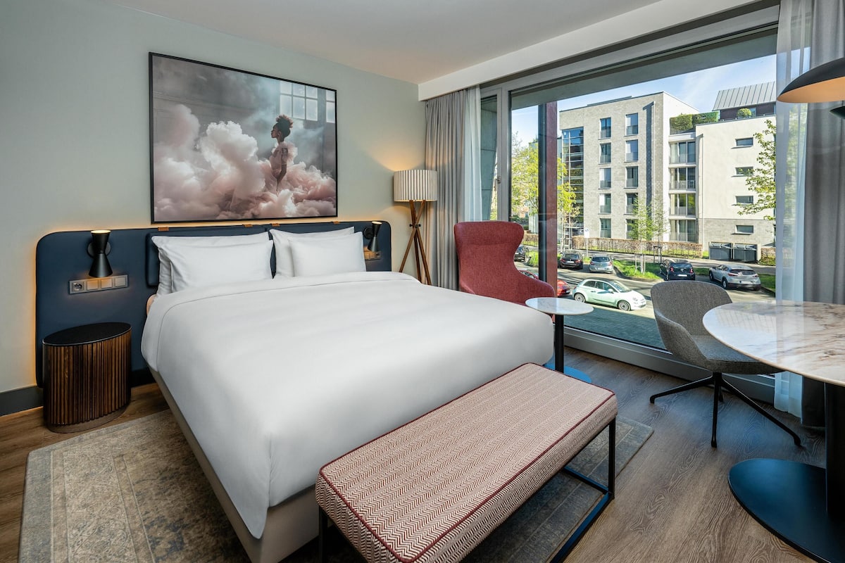 A modern hotel room with a large bed, a bench, a desk, and a chair. There is a large window offering a view of the city.