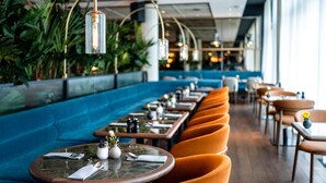 A restaurant with a long blue bench seating area, round tables with orange chairs, and hanging light fixtures. - Radisson Blu Media Harbour Hotel, Düsseldorf (Düsseldorf)
