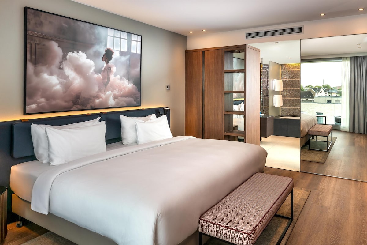A modern hotel room with a large bed, a bench, a large window, and a view of the city.