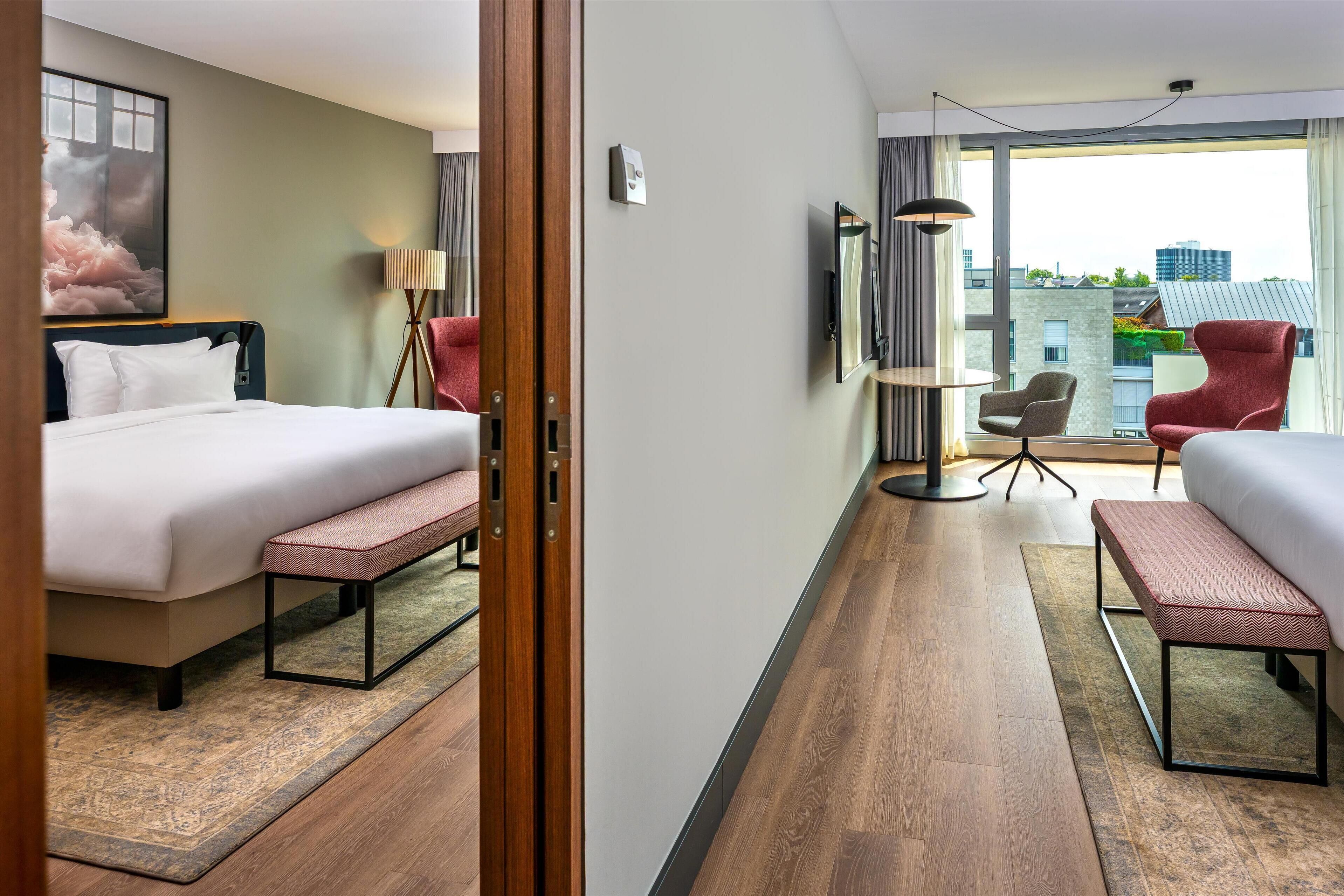 Een hotelkamer met een bed, een bankje, een bureau, een stoel en een groot raam met uitzicht op de stad.